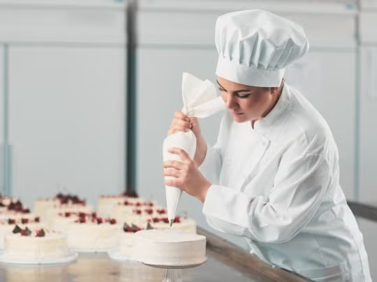 Repostera vestida con uniforme blanco y gorro decora un pastel con una manga pastelera en una cocina