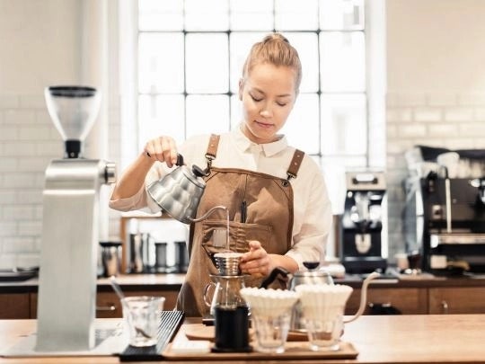 Una mujer barista con delantal vierte agua caliente en un gotero de café en una cafetería moderna.