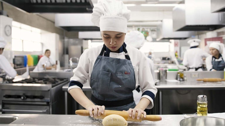 Joven chef del Programa YOCUTA usa rodillo para amasar en una cocina comercial junto a otros estudiantes