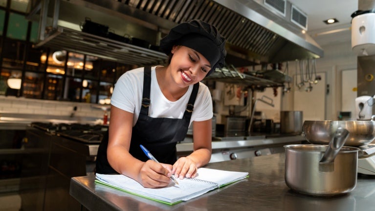 En una cocina comercial, joven estudiante del programa YOCUTA toma notas en su libreta sobre