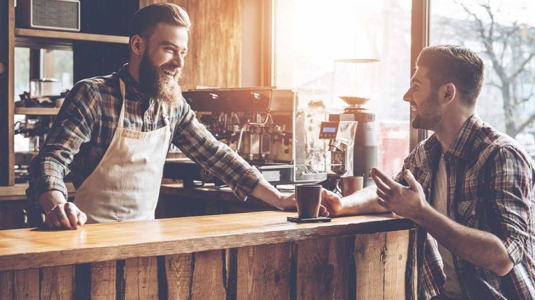 Barista atiende en su cafetería a un cliente sentado a la barra que sostiene una taza de café