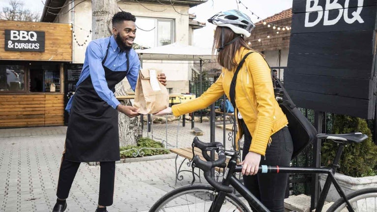 Empleado de restaurante entrega a una clienta que va en bicicleta un pedido a través de take away 