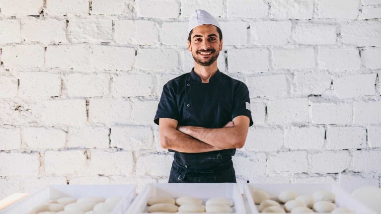 Emprendedor gastronómico sonriendo con los brazos cruzados frente a cestas de masas para panadería