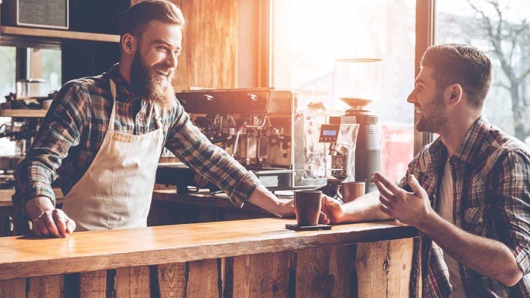 Barista conversa animadamente con cliente sentado a la barra de su negocio que sostiene una taza de café