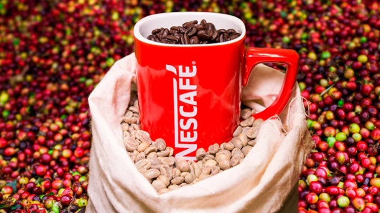 Taza roja llena de café en grano NESCAFÉ® en una bolsa con café verde y rodeado de cerezas