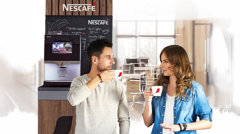 Dos compañeros de trabajo disfrutan de una taza de café en la oficina, al fondo una máquina NESCAFÉ®