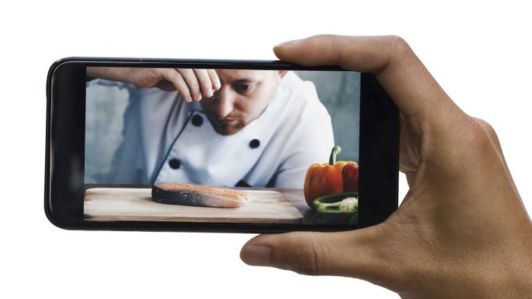 Mano sostiene un smartphone en el que se proyecta un chef sazonando un filete de pescado