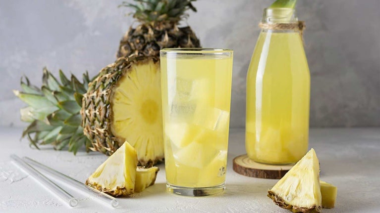 Vaso y botella de cristal de agua saborizada de piña, al fondo está la fruta y los trozos