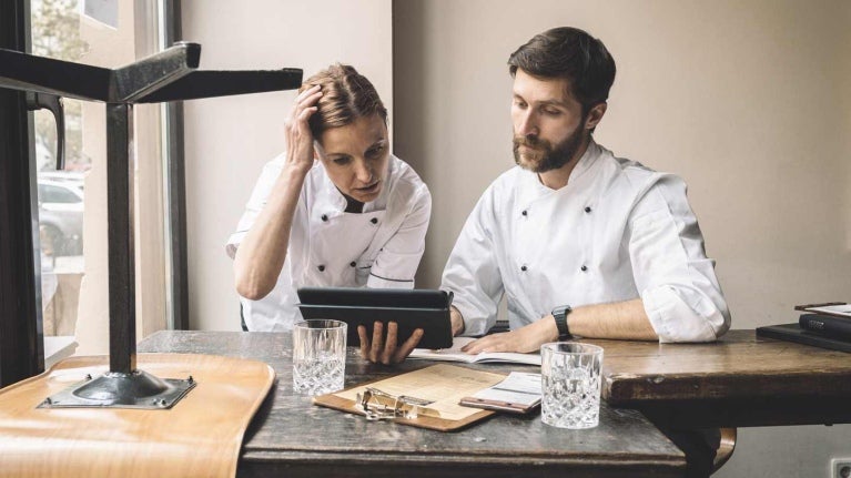 Dos chefs observan una tablet sentados a la mesa donde hay vasos, libretas y tabla legajadora