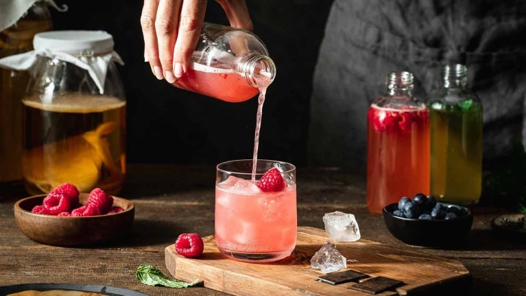 En vaso con hielo y frambuesas mixólogo vierte un líquido de una botella, al lado otras bebidas y frutas