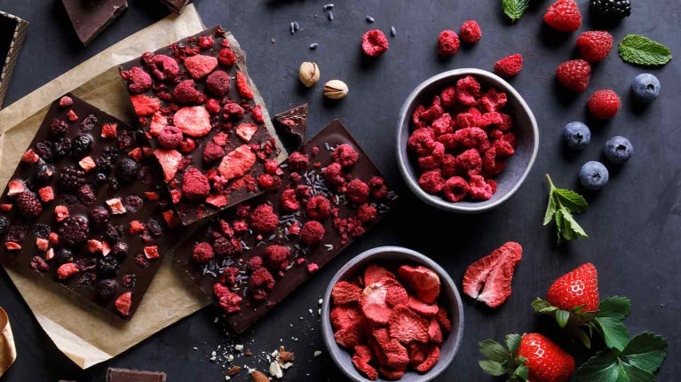 Tabletas de chocolate con frambuesas, arándanos y fresas liofilizadas, al lado las frutas frescas