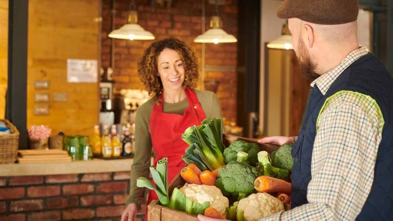 Dueña de un negocio recibe a su proveedor de alimentos local quien lleva una caja con vegetales frescos