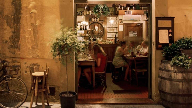 Fachada de una hostería con comensales al fondo sentados a la mesa y afuera sillas, plantas y bicicleta