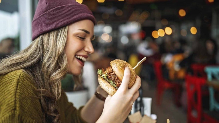 Mujer observa una hamburguesa que sostiene en sus manos, al lado bebida y otros aperitivos