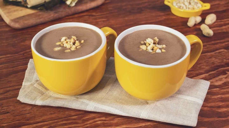 Dos tazas amarillas de chocolate caliente coronadas con maní, al fondo hallacas sobre una tabla