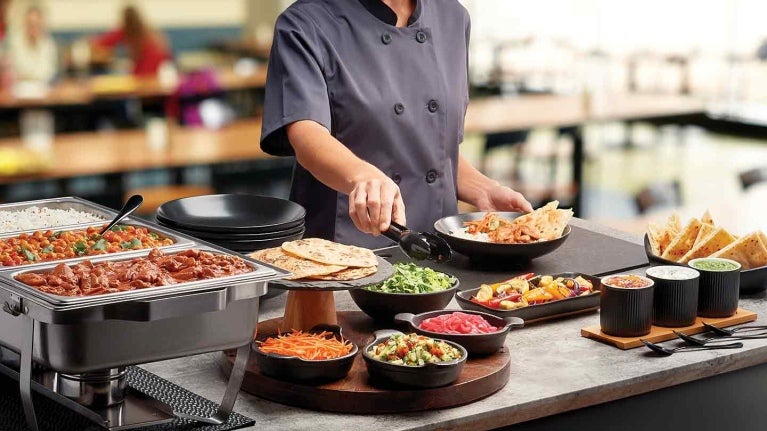 Chef frente de una mesa de buffet sostiene unas pizas para tomar la ensalada de una taza