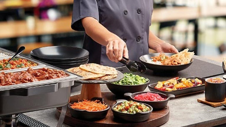 Chef frente de una mesa de buffet sostiene unas pizas para tomar la ensalada de una taza