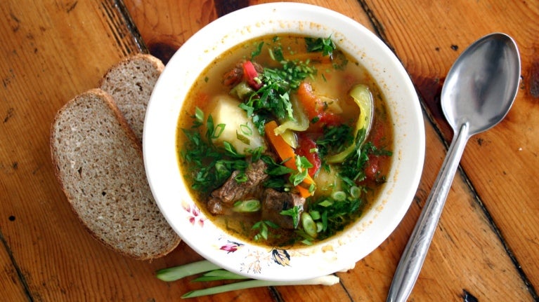 Plato de sopa casera con verduras, carne y pan sobre mesa de madera representando la cocina de soda.