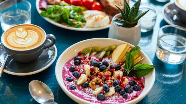 Smoothie bowl decorado con frutas frescas, acompañado de un café latte y un desayuno variado en una mesa.