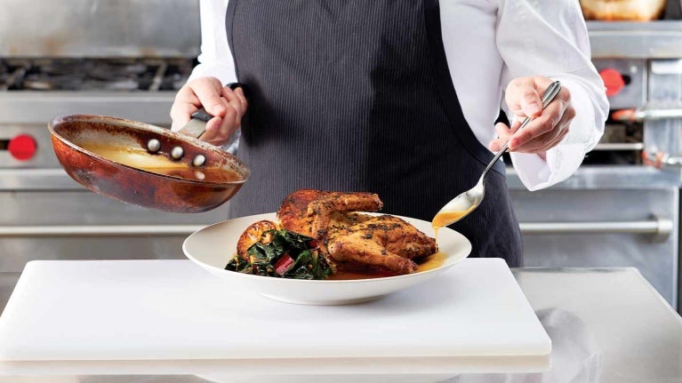 Chef adereza pollo servido en un plato sobre el mesón de una cocina, en otra mano tiene una sartén