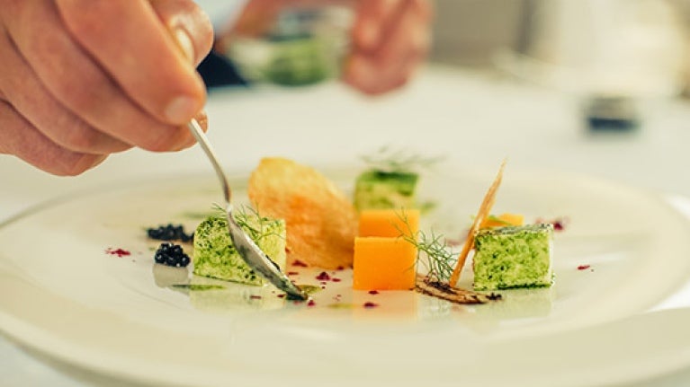 Mano de chef decora con aderezo un plato blanco de vegetales en cubos