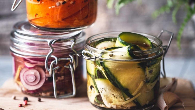 Tres recipientes de cristal con cebolla, pepino y zanahoritas en conserva sobre una tabla rodeada de ajos