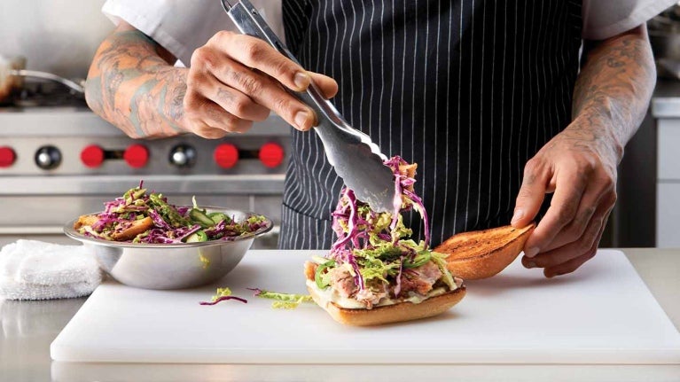 En una cocina comercial, chef agrega ensalada con pinzas al pan sobre una tabla de cortar