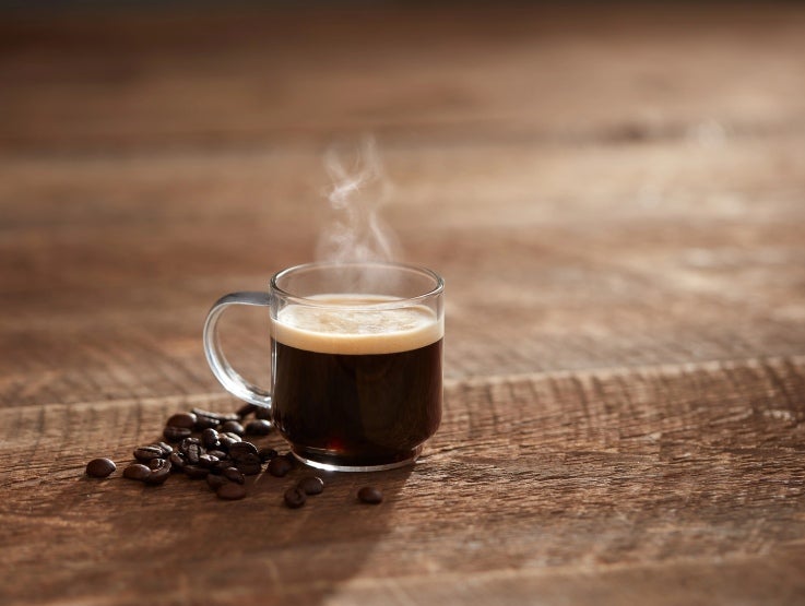 Sobre una mesa de madera, una taza café humeantes de NESCAFÉ® Professional, rodeada de granos tostados