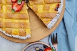 Tarta de ricotta y manjar Nestlé decorado con frutillas y borde de coco rallado junto a un plato con una porción de la tarta